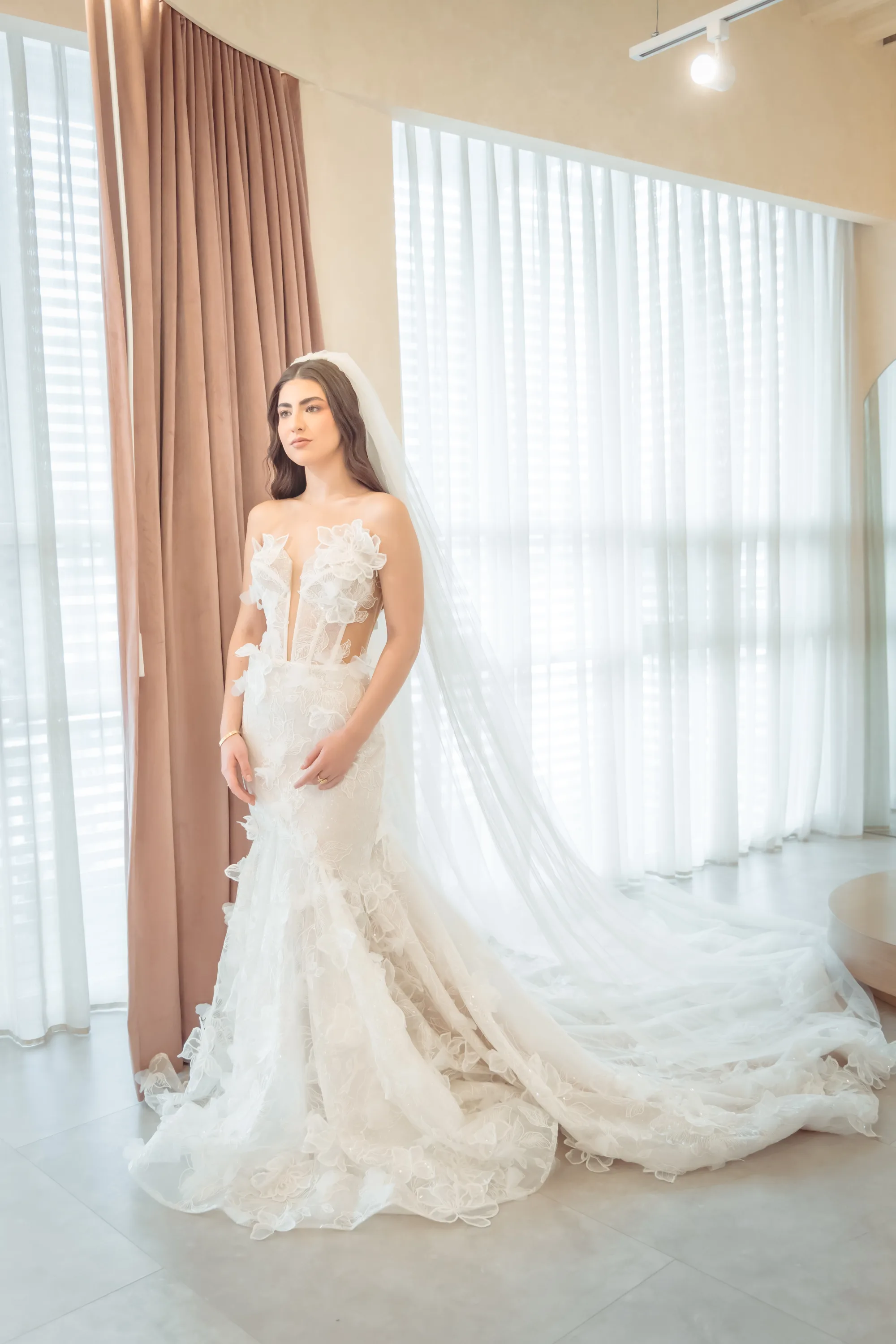 Novia con vestido sirena de encaje floral y velo, frente a cortina y ventanales, fotografía real de Rabling Bridal